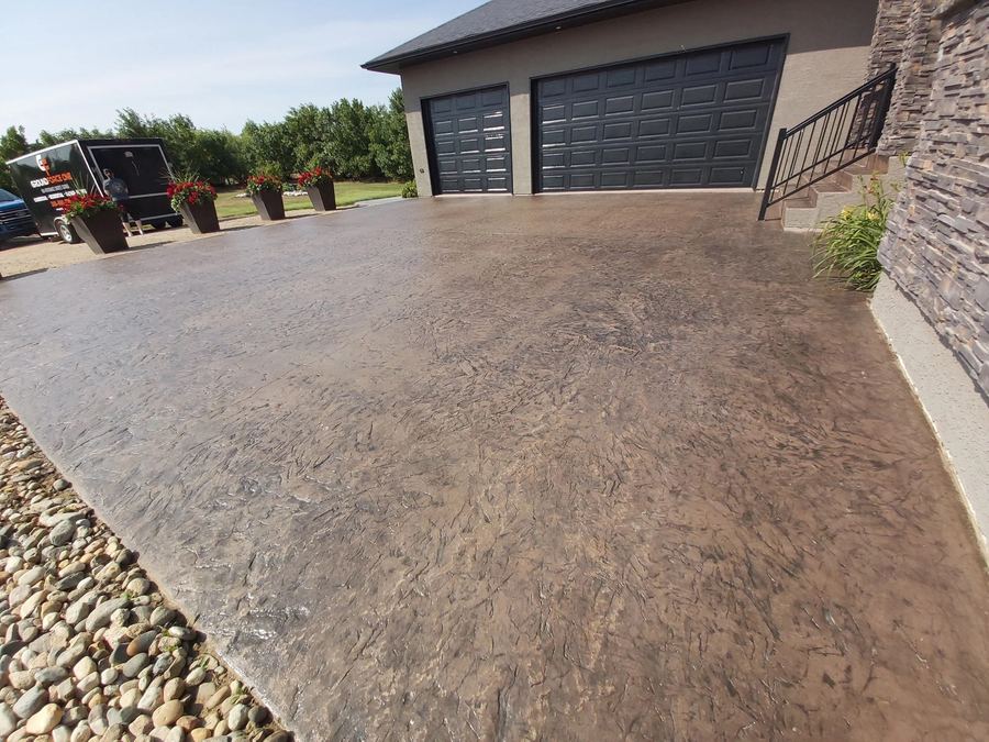 Deep brown sealed driveway bordered by river rock next to a double garage